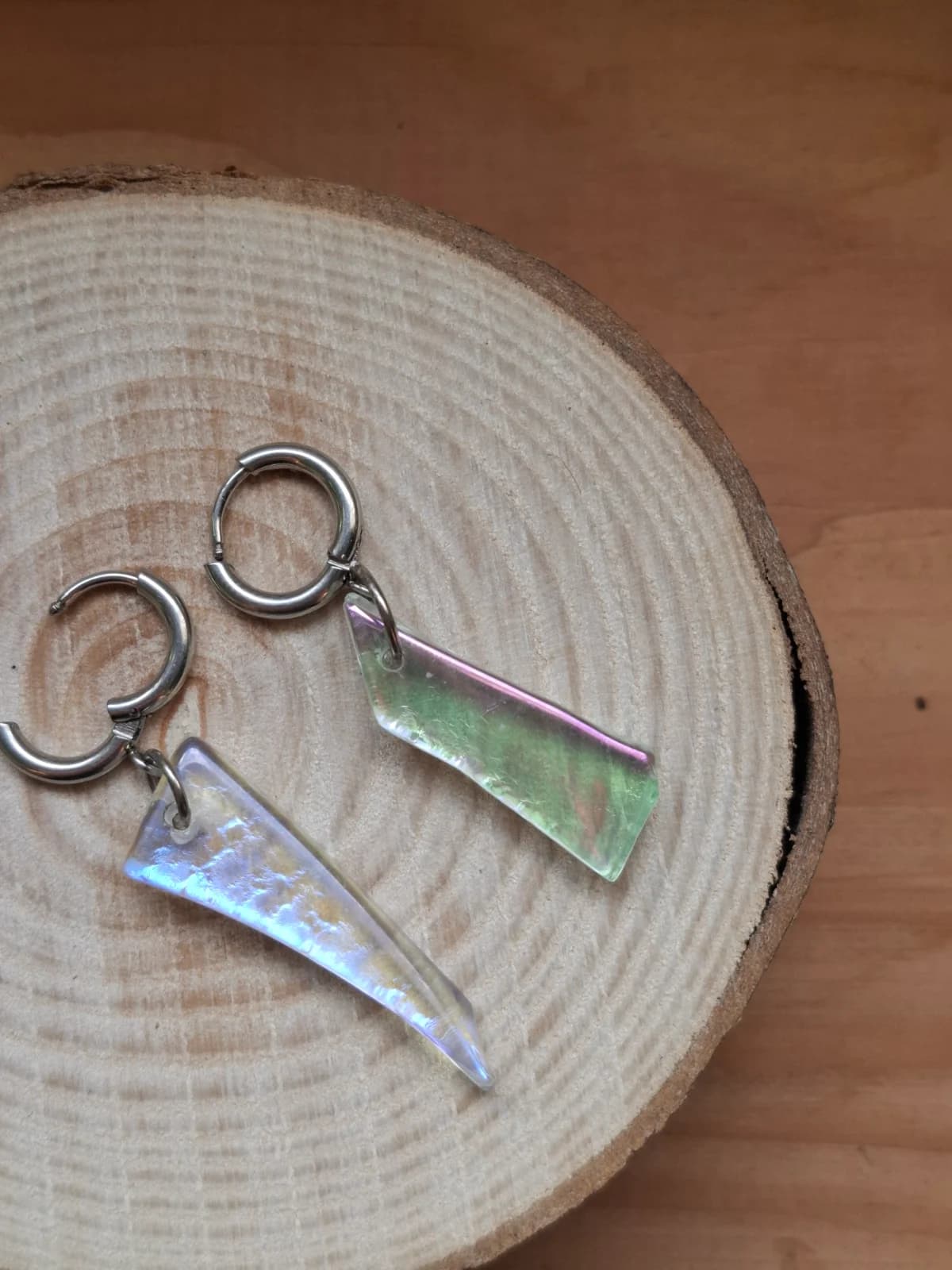 Boucles d'oreilles pendantes en verre holographique aux reflets verts et mauves sur un rondin de bois.