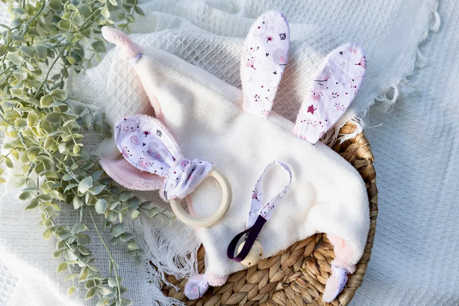 Doudou plat oreilles de lapin écru et rose, anneau de dentition et attache-sucette dans un panier tressé