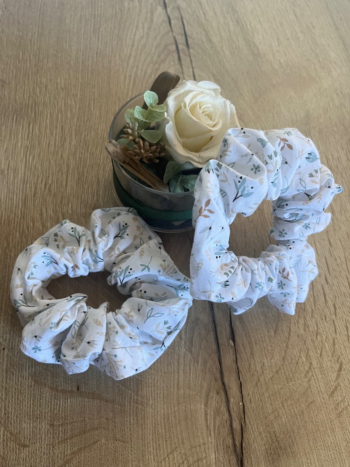Deux chouchous en tissu clair à motif de feuilles posés sur une table en bois à côté d'une rose blanche.