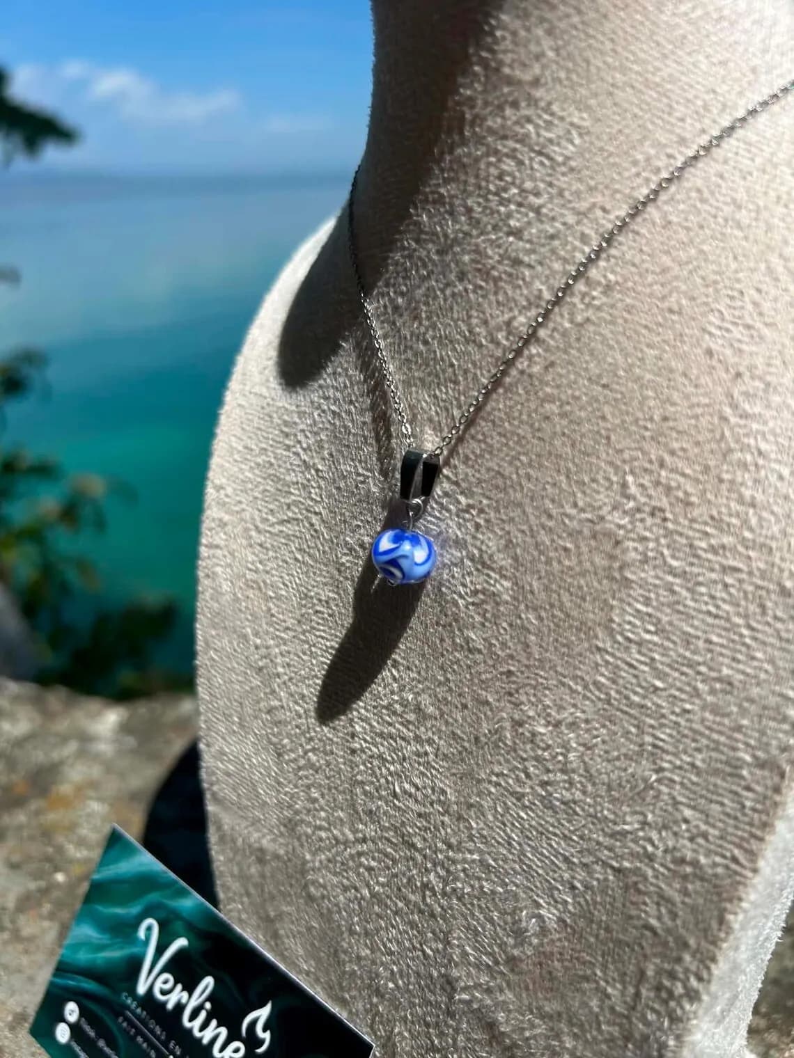 Collier avec perle en verre filé bleu et blanc sur un buste d'exposition devant la mer