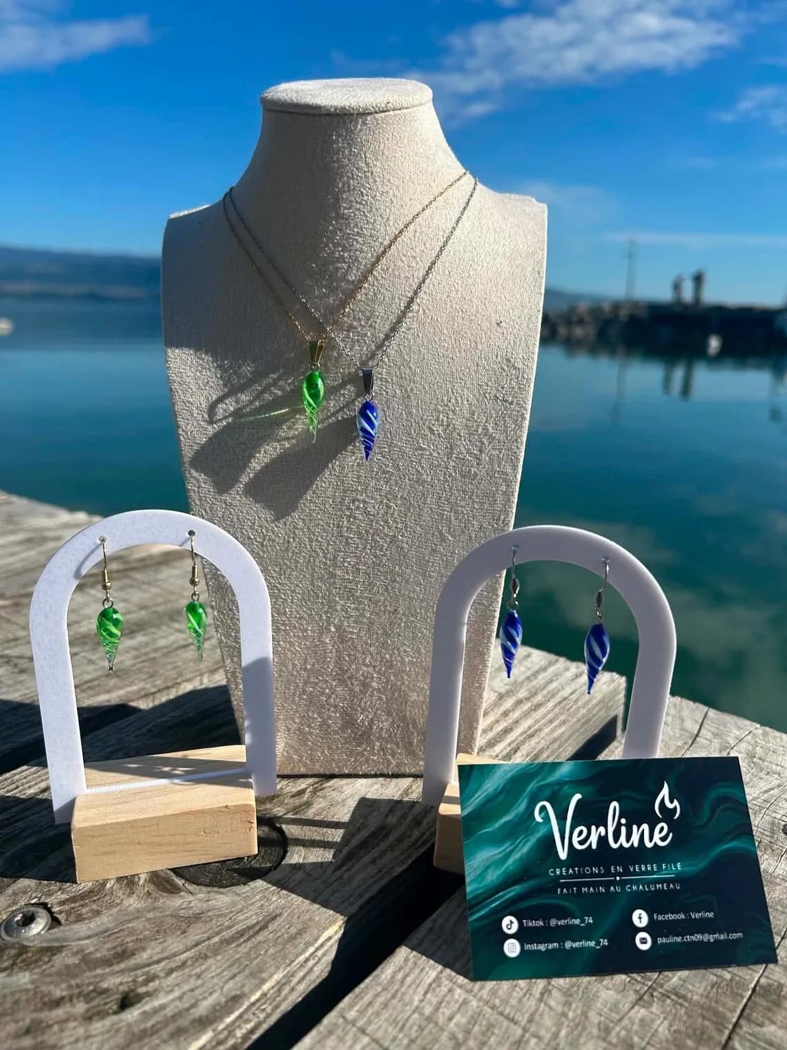 Pendentifs et boucles d'oreilles en gouttes de verre torsadé bleu et vert sur présentoirs face à l'eau.