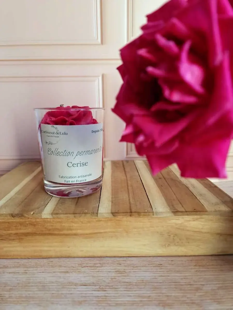 Bougie parfumée Cerise en pot de verre sur un socle en bois, devant une fleur rose foncé